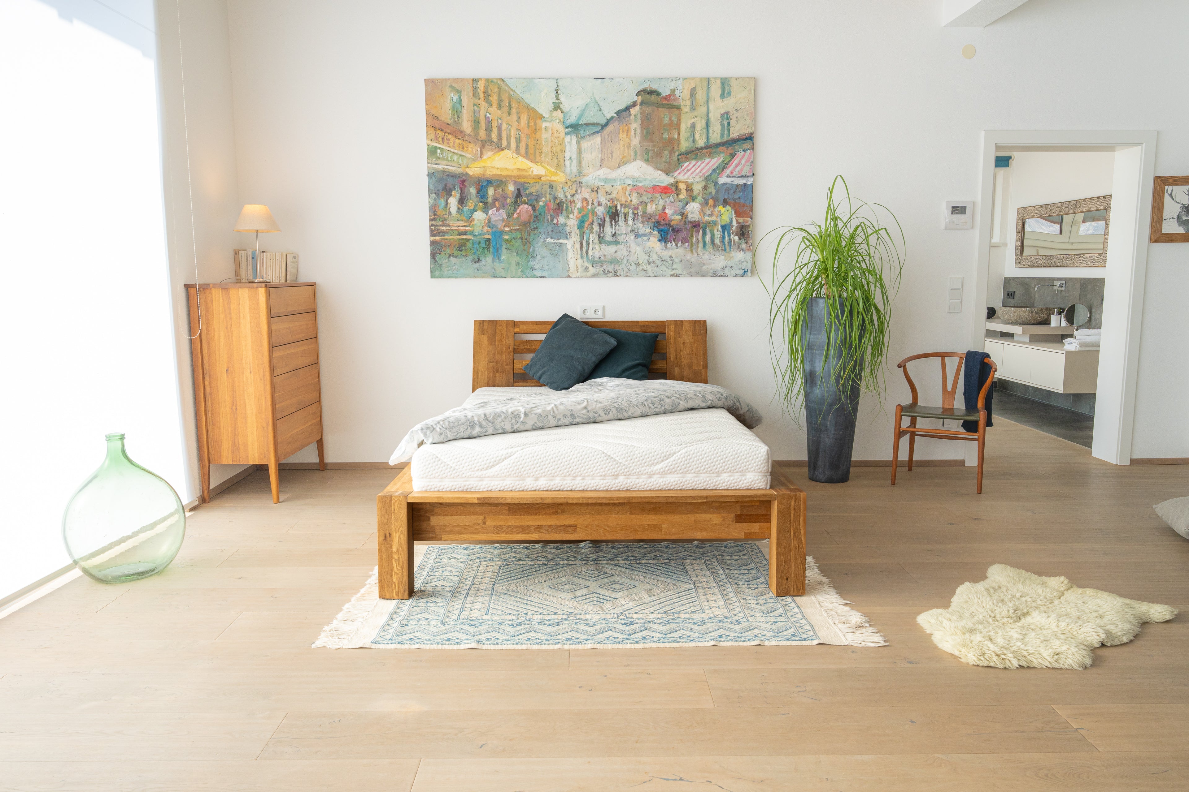 Modernes Schlafzimmer mit einem Holzbett, auf dem die FleXleep-Matratze von xleep liegt. Die weiße Matratze mit ergonomischem Konturschnitt ist stilvoll in Szene gesetzt, ergänzt durch weiche Bettwaren und ein gemütliches Ambiente mit Holzmöbeln, einem großen Gemälde und Pflanzen.
